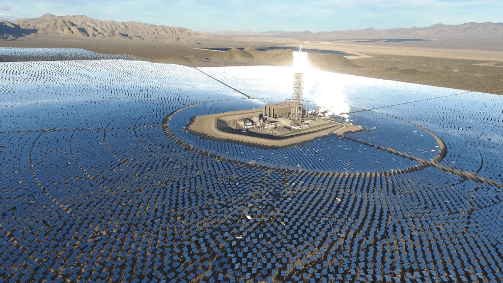 Aerial view of the Ivanpah Solar Power Facility with solar panels arranged in concentric circles around a central tower, set against a desert landscape.