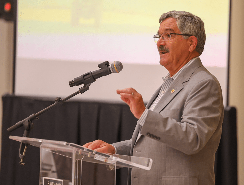 A man in a light gray suit speaks at a podium with a microphone during a presentation, addressing an audience in a conference setting.